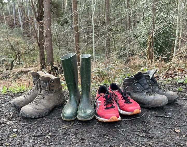 Walking boots, wellies and walking shoes on a muddy path with forest in the background.