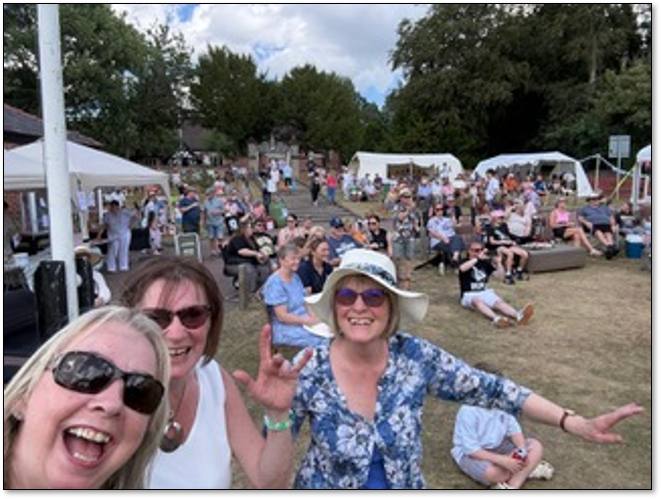Three ladies celebrating at the Festival with many people watching the stage from the banking behind them