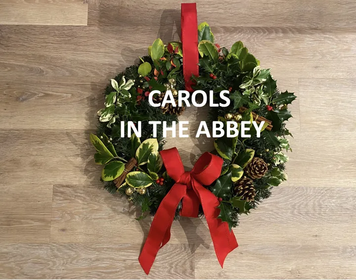 A Christmas wreath with holly and red ribbons and the words "Carols in the Abbey" written over the centre.